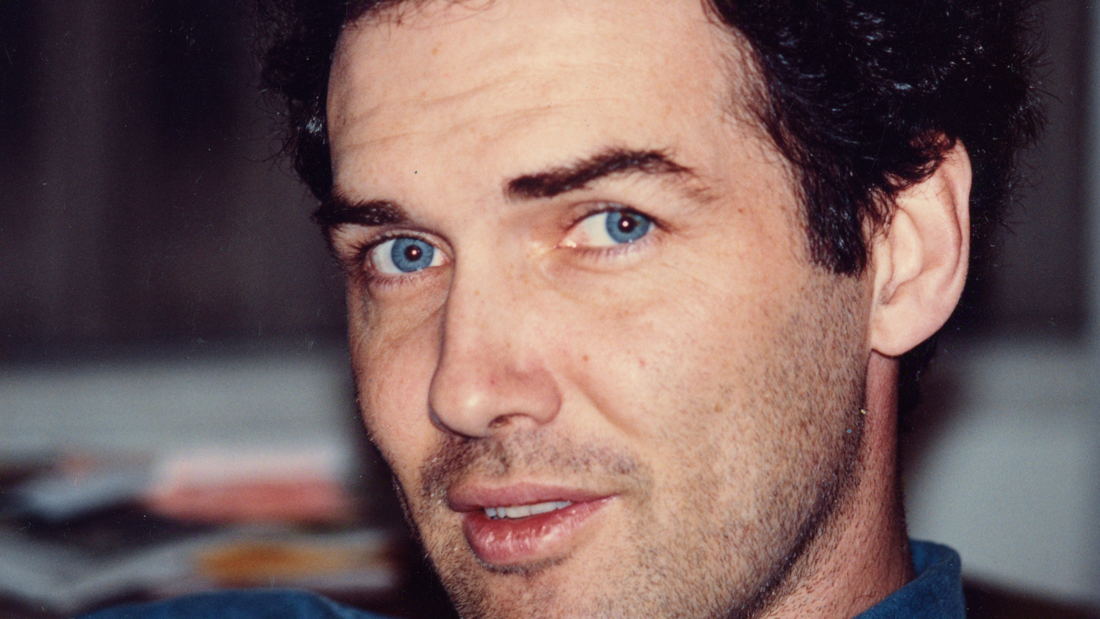 "Man with blue eyes and dark curly hair looking at the camera indoors, with blurred background and casual setting."